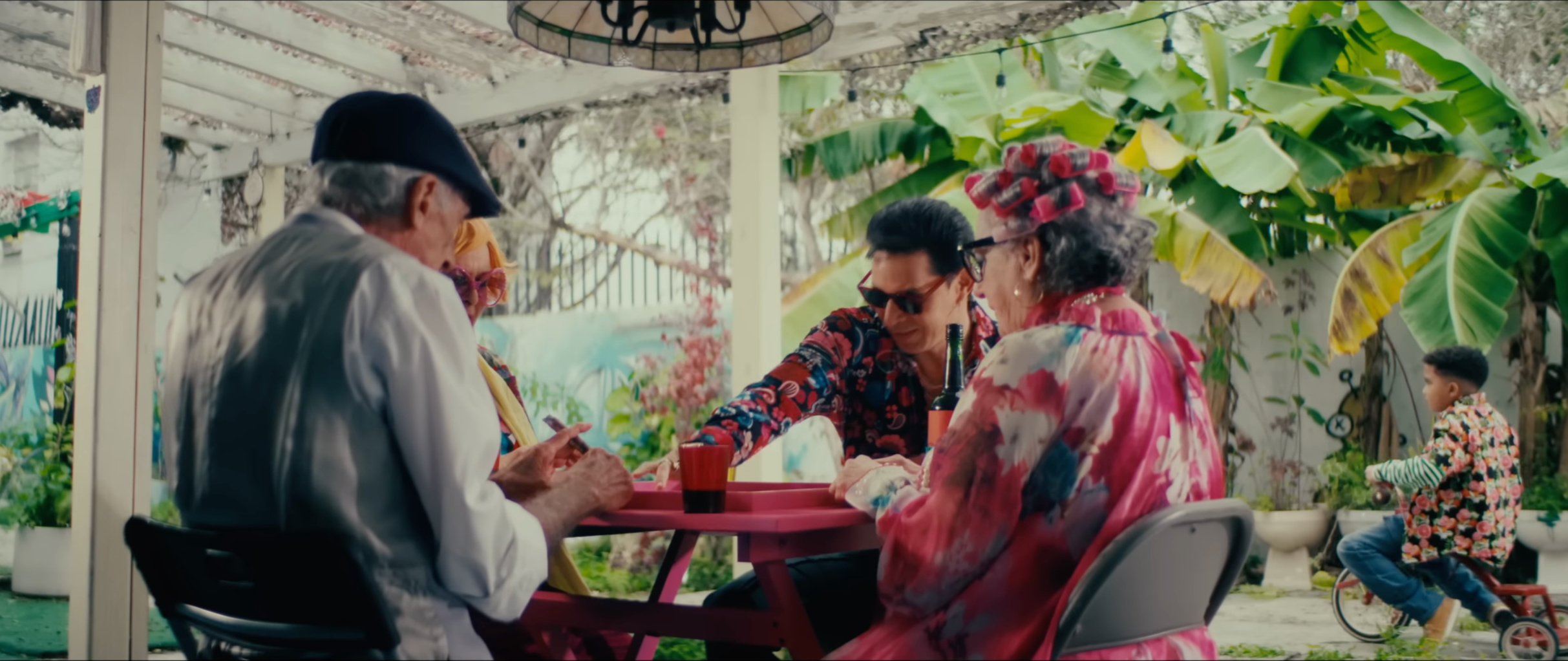 Group of four older adults and one young child sitting at a pink outdoor table enjoying drinks and conversation in a lush garden with banana plants, a clock, and a child riding a bicycle in the background.
