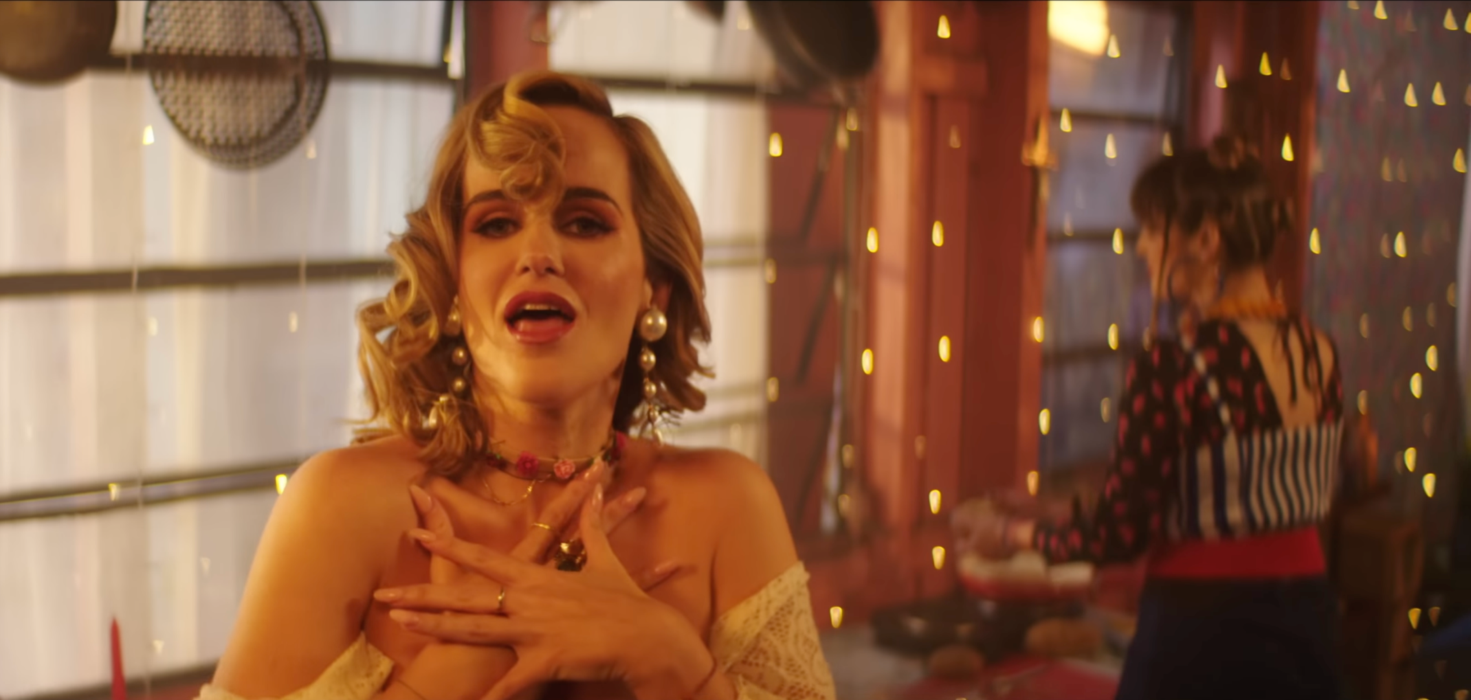 A woman with vintage style hair and makeup, wearing large pearl earrings and colorful jewelry, holding her hands to her chest in an emotional moment, in a warmly lit room with string lights.