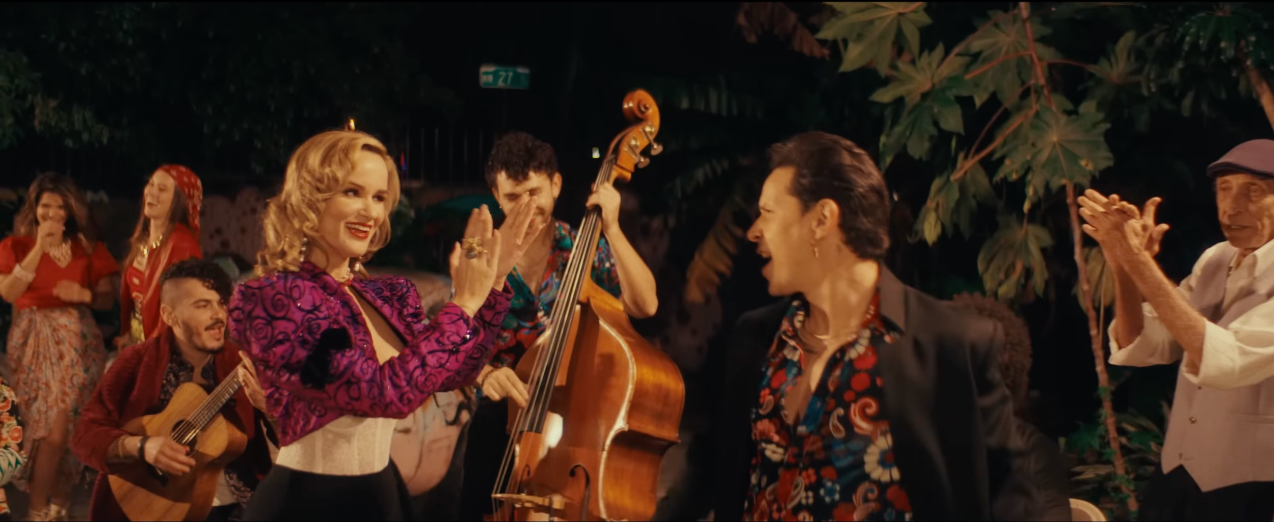 A group of people at a lively outdoor party at night, with some playing musical instruments and others dancing and clapping, surrounded by tropical plants.