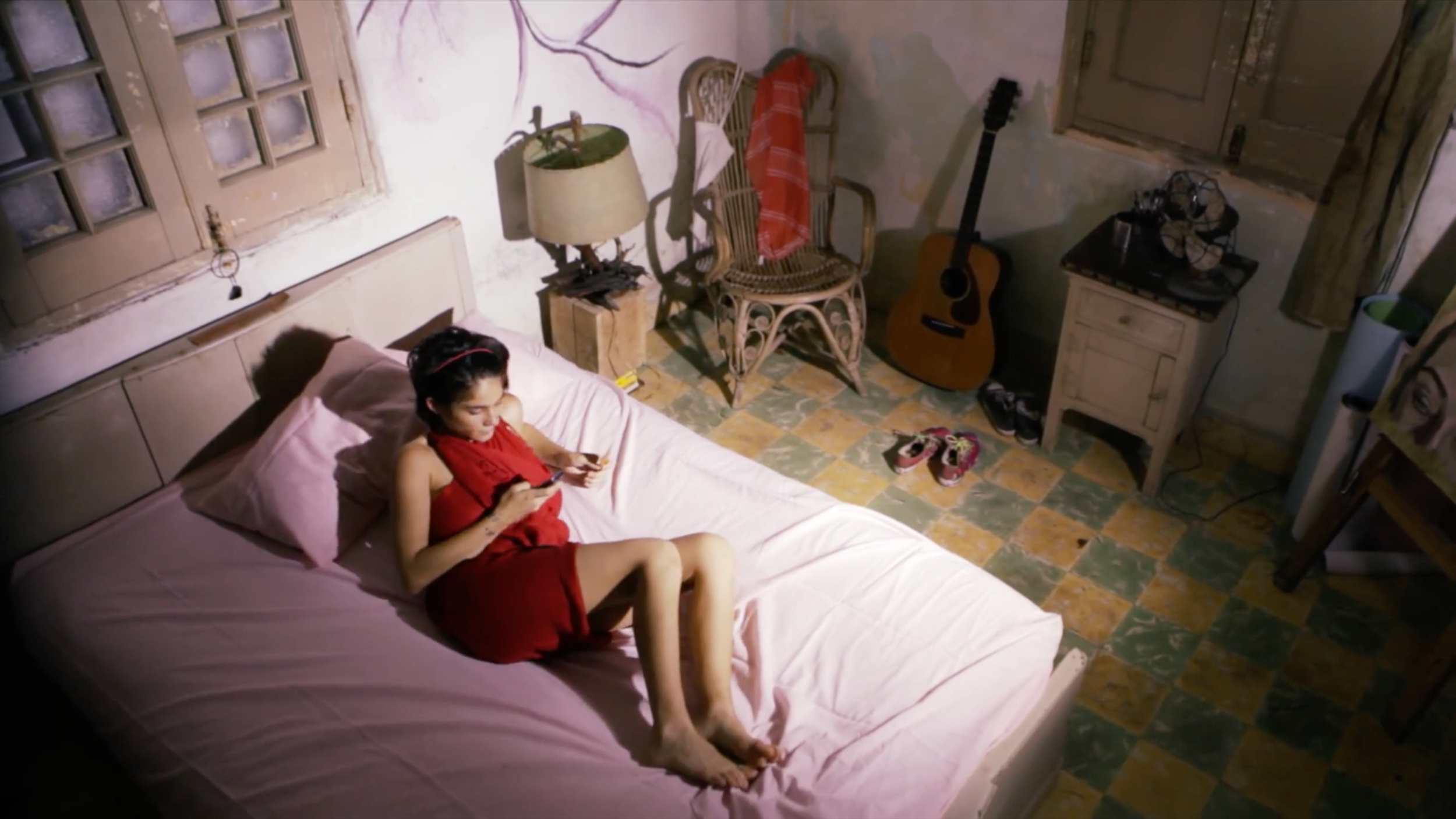 Patricia Machin Portfolio Designer Art director Miami A young woman in a red dress lying on a bed, looking at her phone in a dimly lit, cozy bedroom with a window, a wicker chair, a guitar, shoes on the floor, and various personal items.