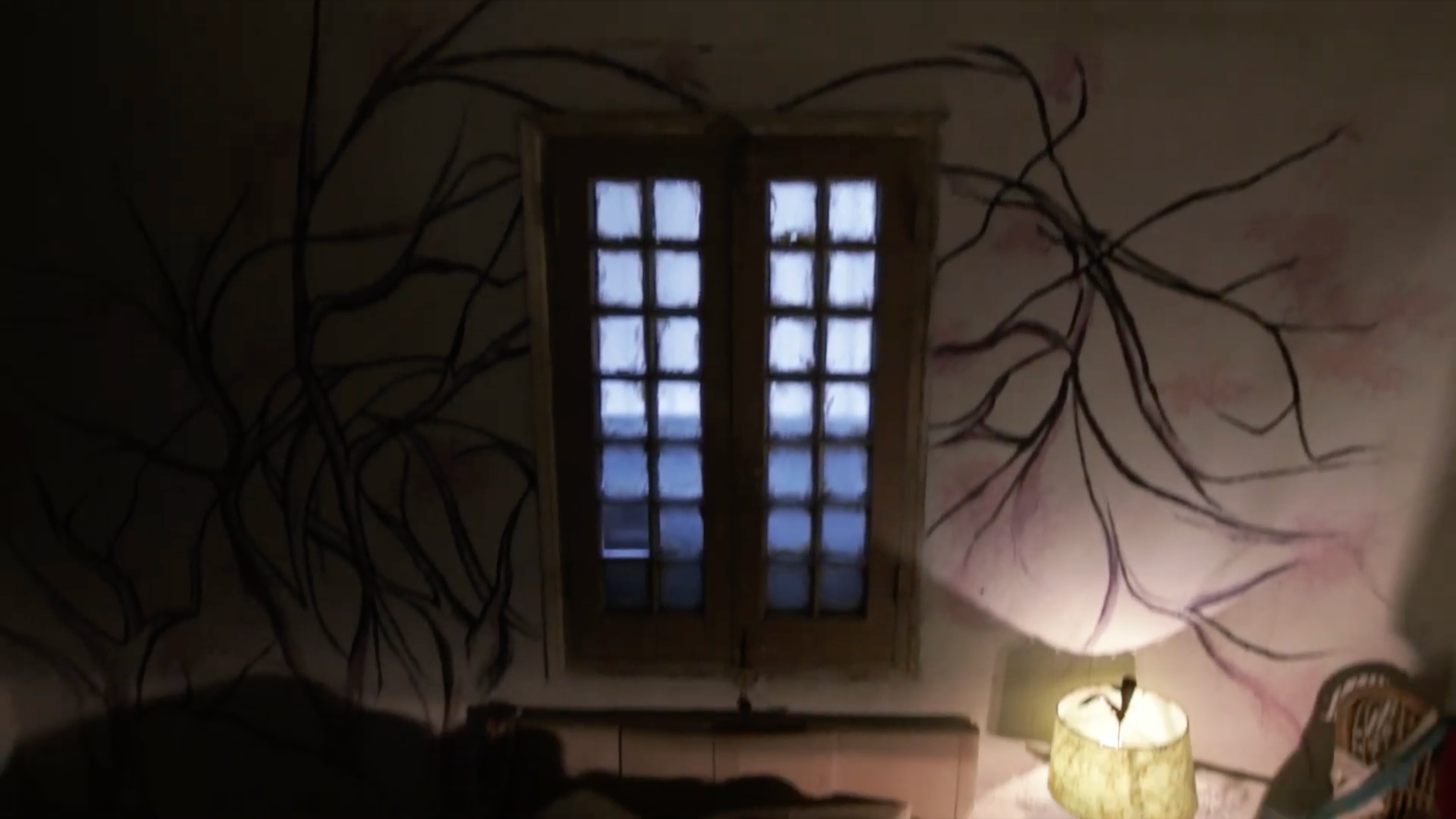 Patricia Machin Portfolio Designer Art director Miami A dimly lit room with a window divided into small rectangles, a lamp on a table, and a wall with painted tree branches.