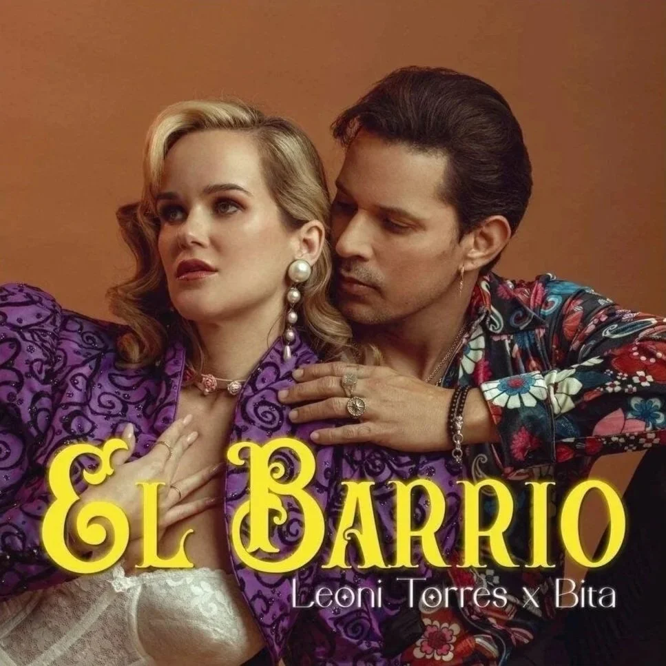 A man and woman pose for a photoshoot with vintage-inspired fashion. The woman has blonde hair styled in waves, wears large pearl earrings, a choker, and a purple embroidered jacket over a lace top. The man has dark, slicked-back hair, wears rings an