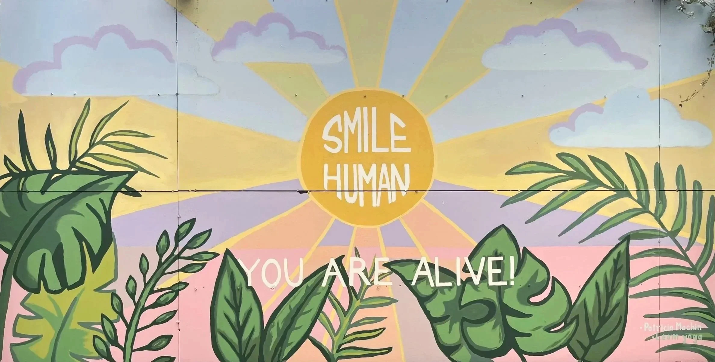 Patricia Machin Portfolio Designer and Art Director Colorful mural with a yellow sun and clouds, featuring green tropical leaves and the message 'Smile Human, You Are Alive!'