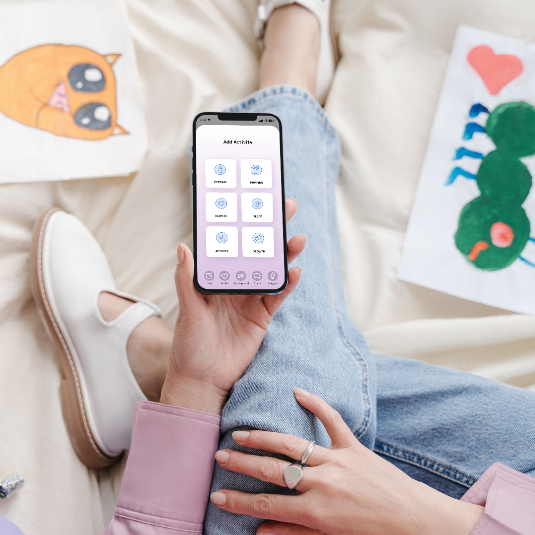 Person sitting on a bed using a smartphone with an app displaying activity options like feeding, purring, dampers, sleep, activity, and growth. The person is wearing jeans, a pink jacket, and white shoes, with artwork of a cartoon face and a caterpillar on the bed.