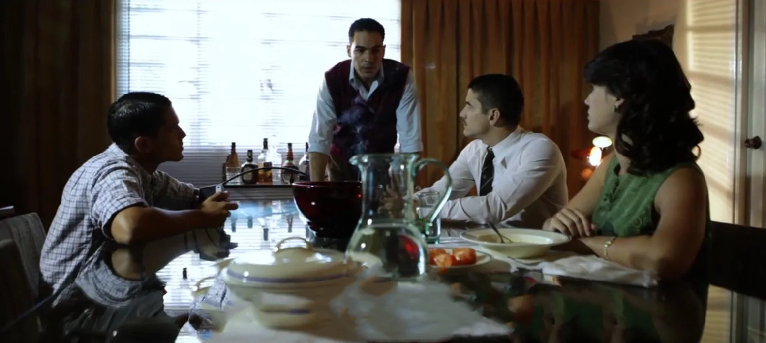 Patricia Machin Portfolio Designer Art director Miami Four people sitting at a dining table in a room with wooden walls, bottles, and a pitcher with food and drinks, engaging in a conversation with a man standing.