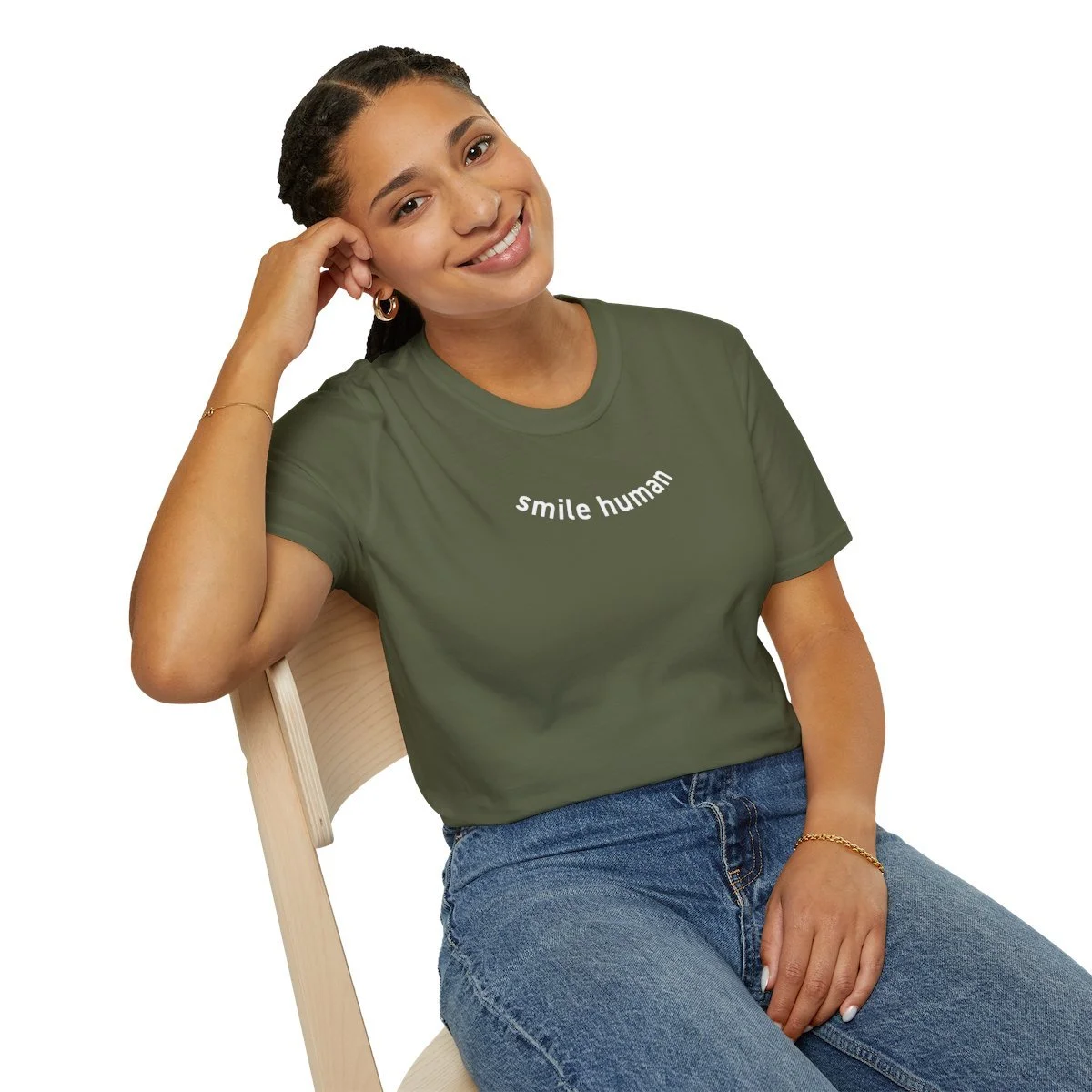 A young woman with braids, smiling and sitting on a light wooden chair. She is wearing a green T-shirt with the words 'smile human' printed on it and blue jeans, accessorized with small hoop earrings and a bracelet.