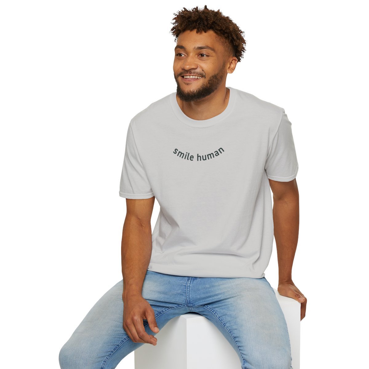 Patricia Machin Miami Portfolio Designer Art Director  A man with curly hair and a beard sitting on a white cube, smiling, wearing a white t-shirt with the words 'smile human' printed on it and light blue jeans.