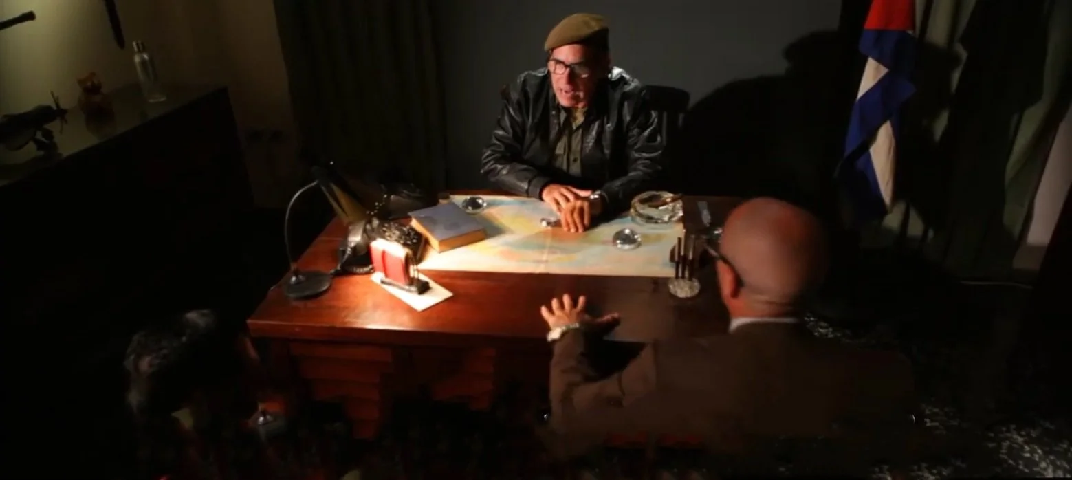 Two men engaged in a conversation across a desk, illuminated by a desk lamp. The man on the phone is seated to the left, and the other man, wearing glasses and a suit, is gesturing with his hands. A map, a book, and a glass of water are on the desk. 