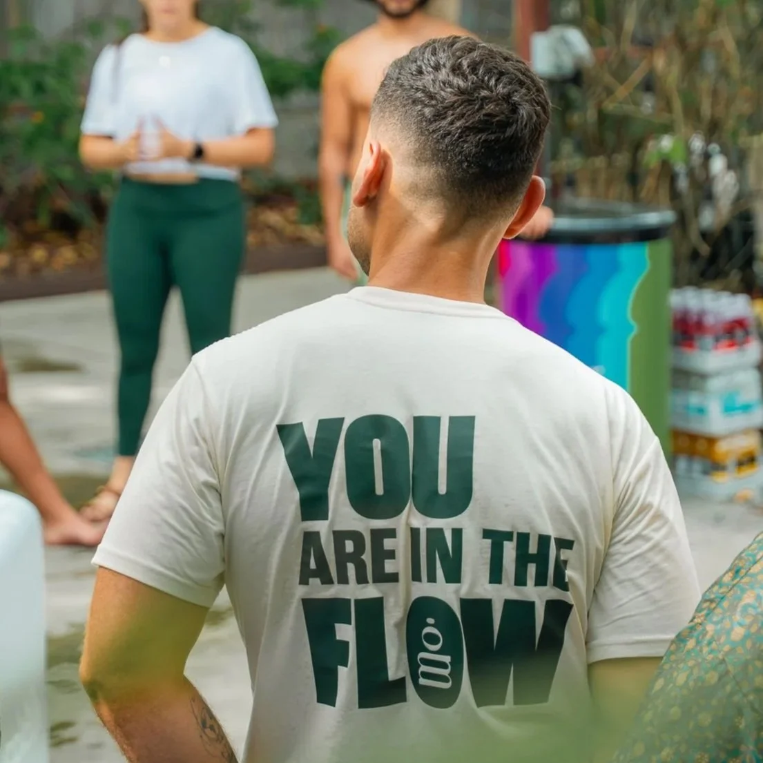 Patricia Machin Portfolio Designer and Art Director Back of a man Leom Wellbeing wearing a white shirt with the text "YOU ARE IN THE FLOW", standing outdoors with people in the background Miami