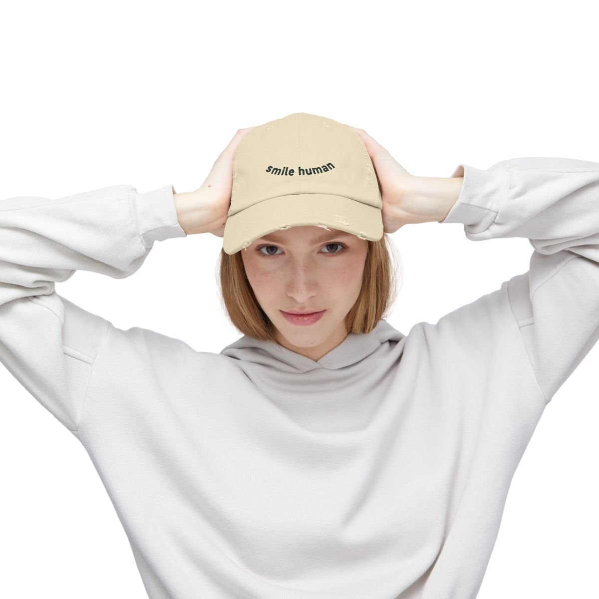 Young woman wearing a beige baseball cap with the words "smile human" printed on it, holding the cap with both hands, dressed in a white sweatshirt, posing against a white background.
