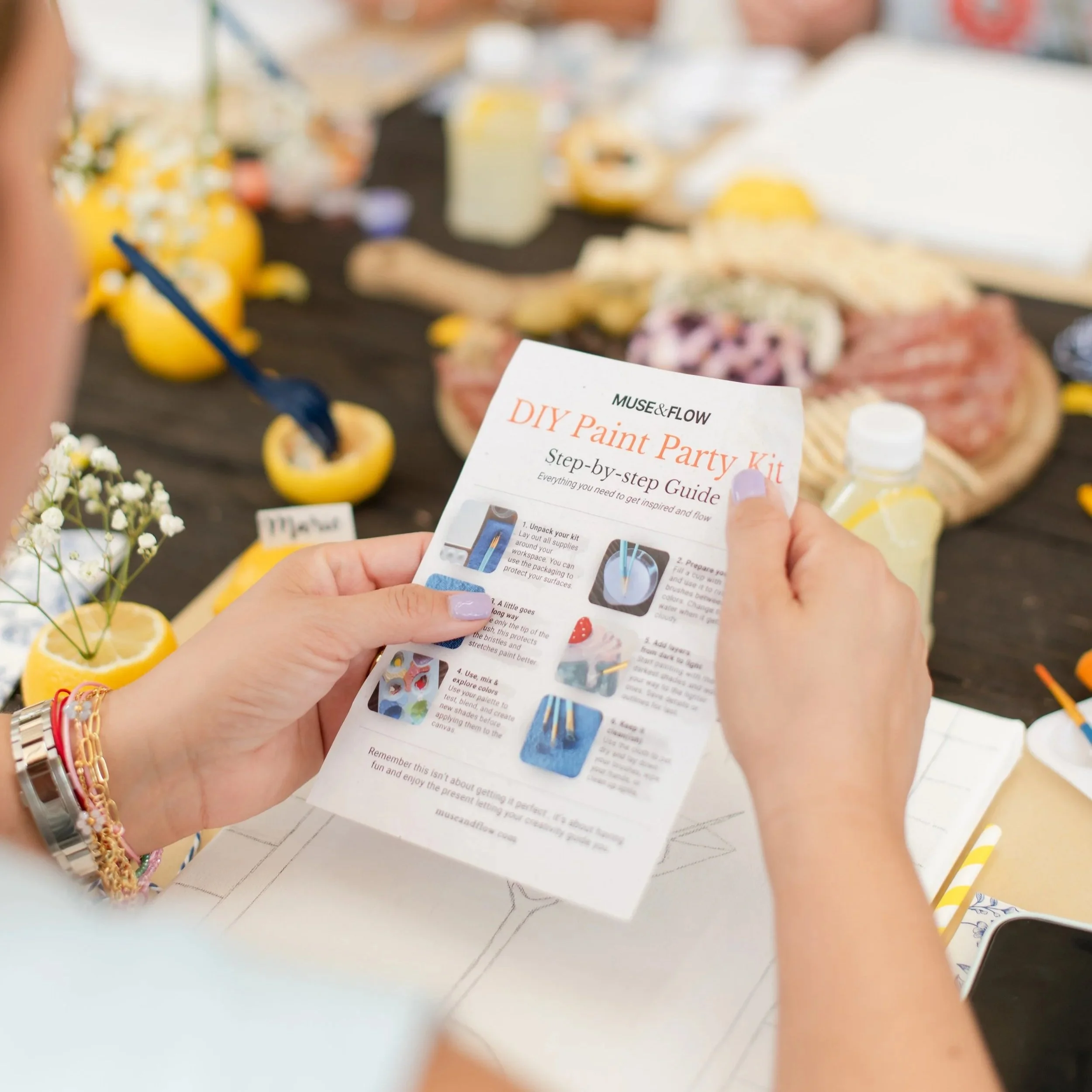 Patricia Machin Portfolio Designer and Art Director Person holding a DIY paint party kit step-by-step guide at a decorated table with lemon and flower decorations, with food and drinks in the background.