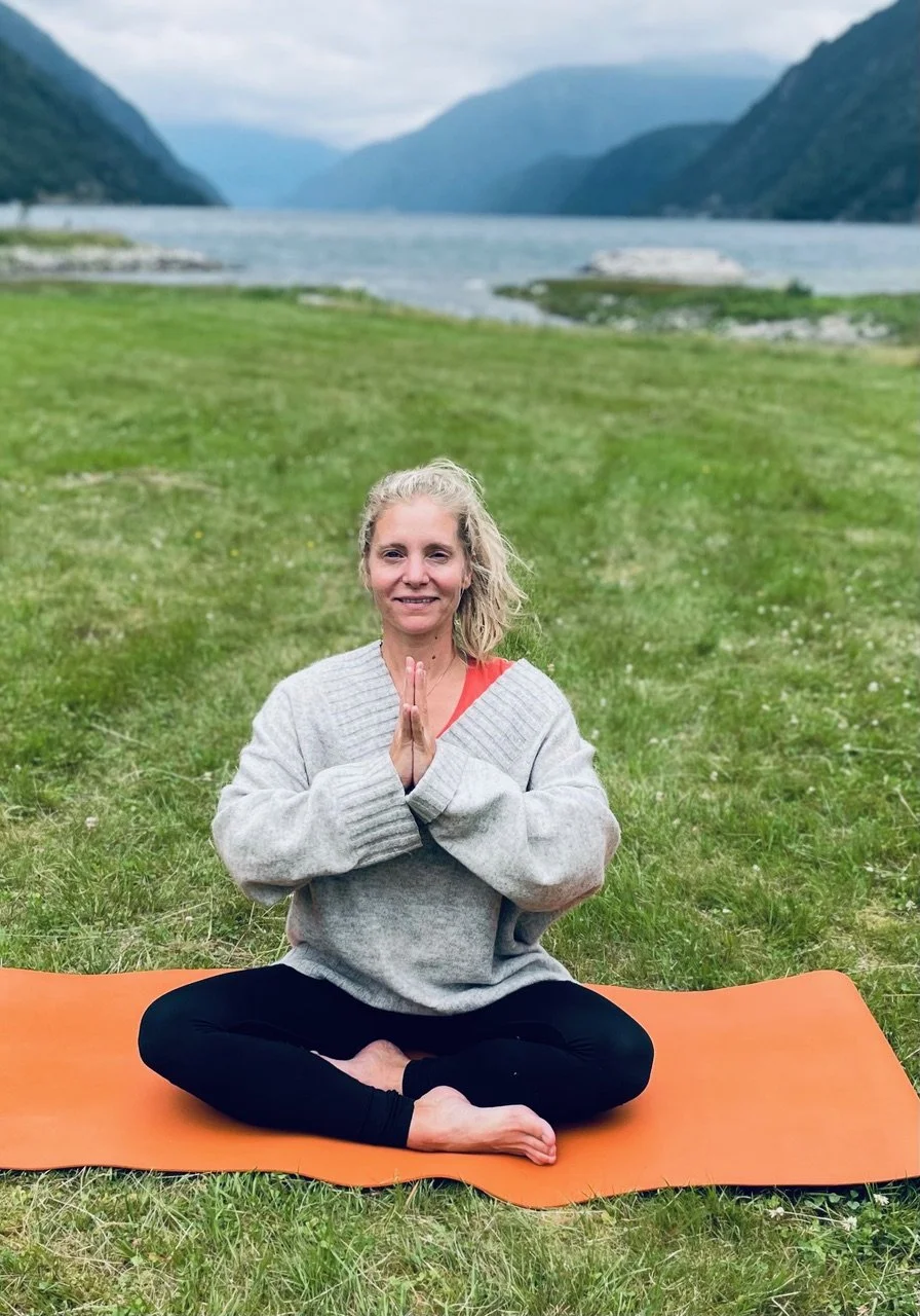 En kvinne sitter i lotusposisjon på en oransje yogamatte på gress, med fjell og innsjø i bakgrunnen.