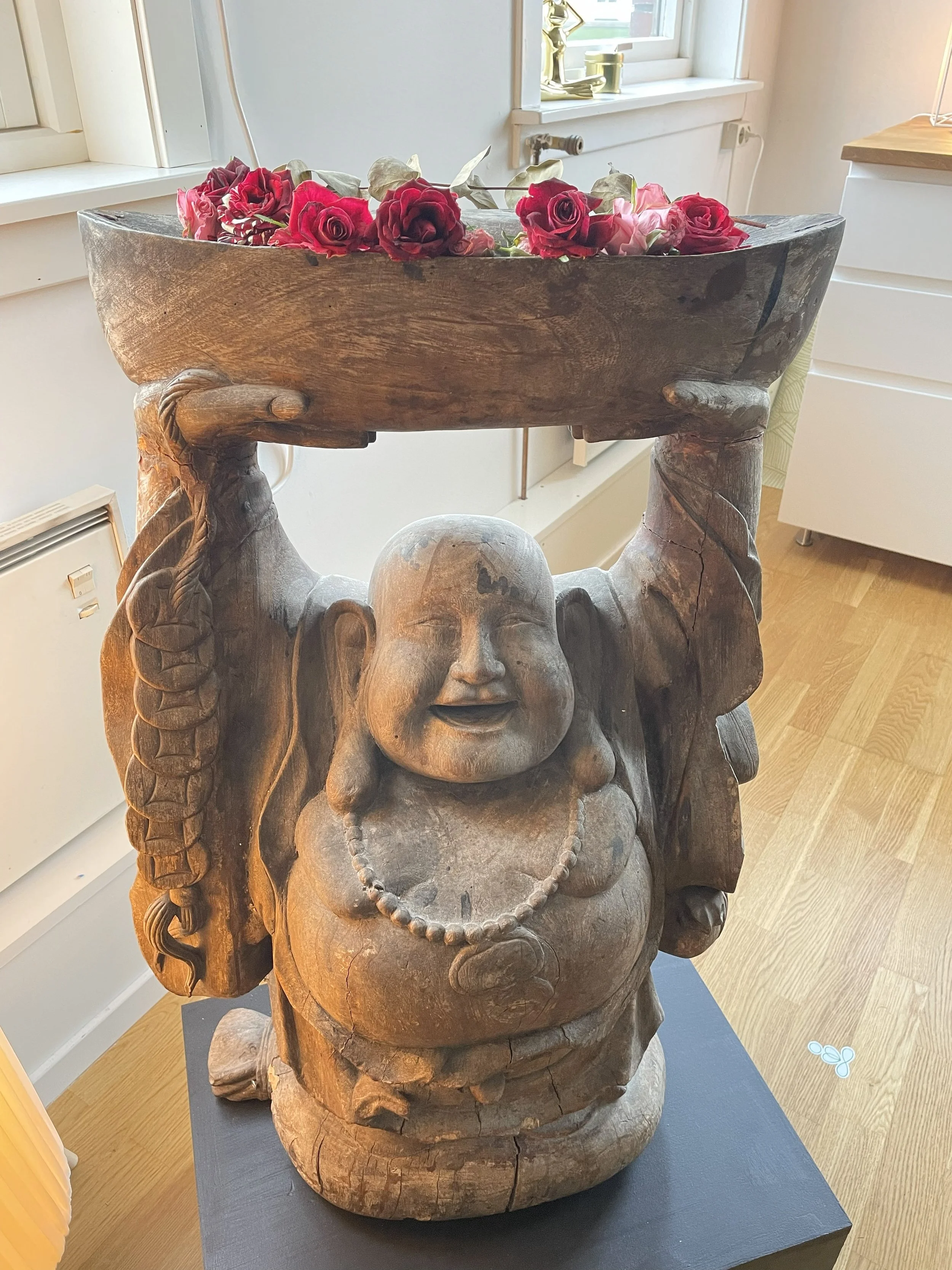 Trefigur av en glad, rund Buddha-munk med store ører, som holder opp en stor blomsterkasse med røde og rosa blomster.