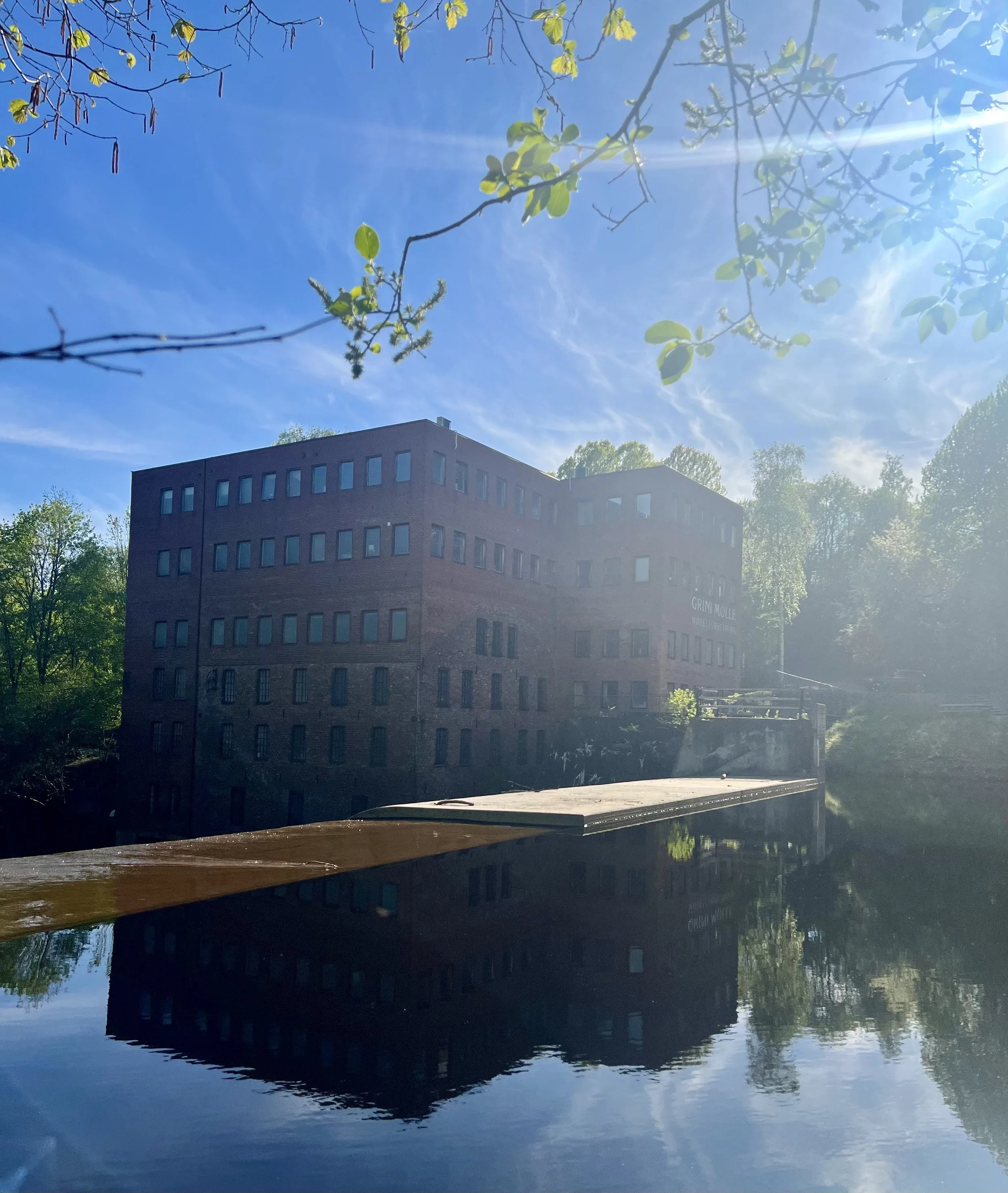 En grav tilgjengelig bygning med flere etasjer og mange vinduer, omgitt av trær, med vann i forgrunnen som reflekterer bygningen og en klar blå himmel med solskinn.