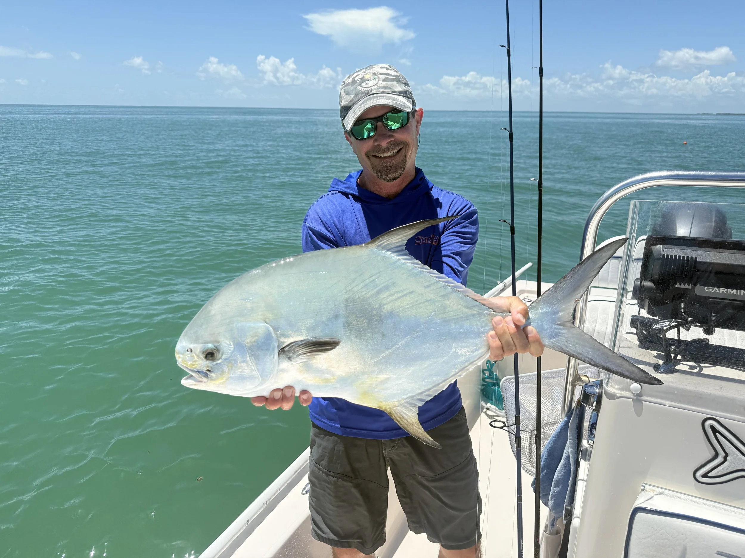 Man on a boat holding a large fish, smiling. He is wearing a gray cap, reflective sunglasses, a blue long-sleeve shirt, and gray shorts. The boat is in the ocean with clear skies and some clouds.