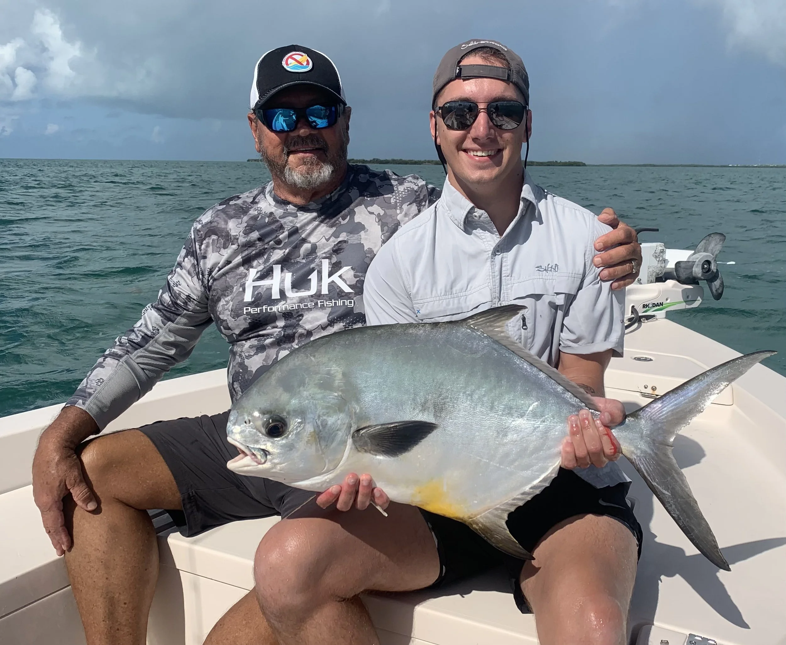 Two men on a boat holding a large fish. One man is older with a beard, wearing sunglasses, a cap, and a camouflage shirt. The other is younger, smiling, wearing sunglasses, a cap backwards, and a light shirt. The background shows the ocean and a clou