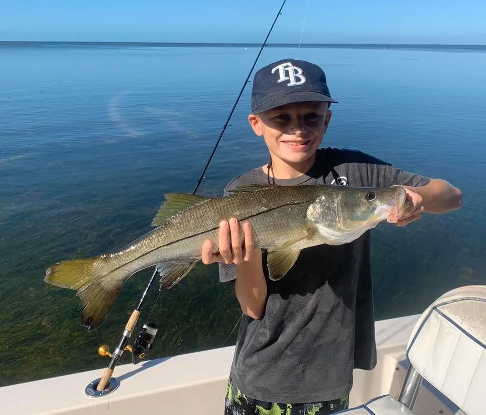 Young boy wearing a dark baseball cap holding a large fish on a boat, with calm water in the background.