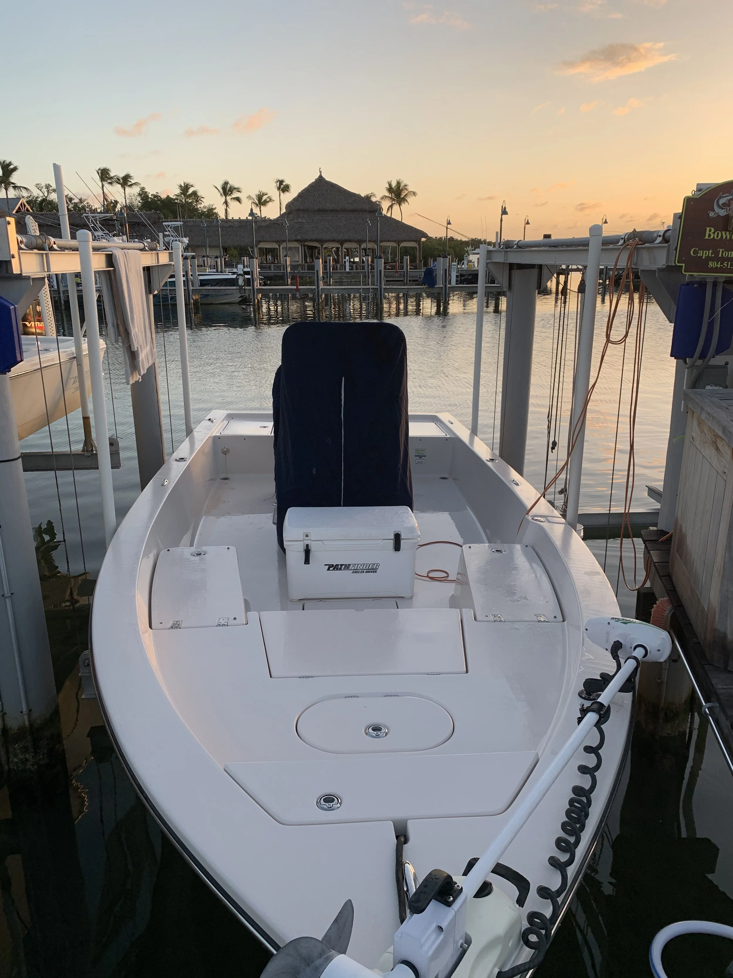 A white fishing boat docked at a marina during sunset, with palm trees and a thatched roof pavilion in the background.