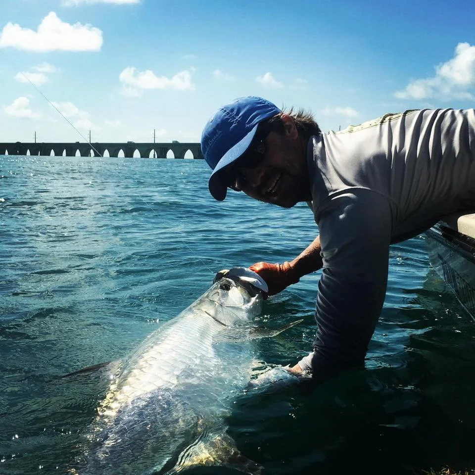 An man in a blue cap and gray shirt is holding a large fish near a body of water, with a bridge in the background and a partly cloudy sky.