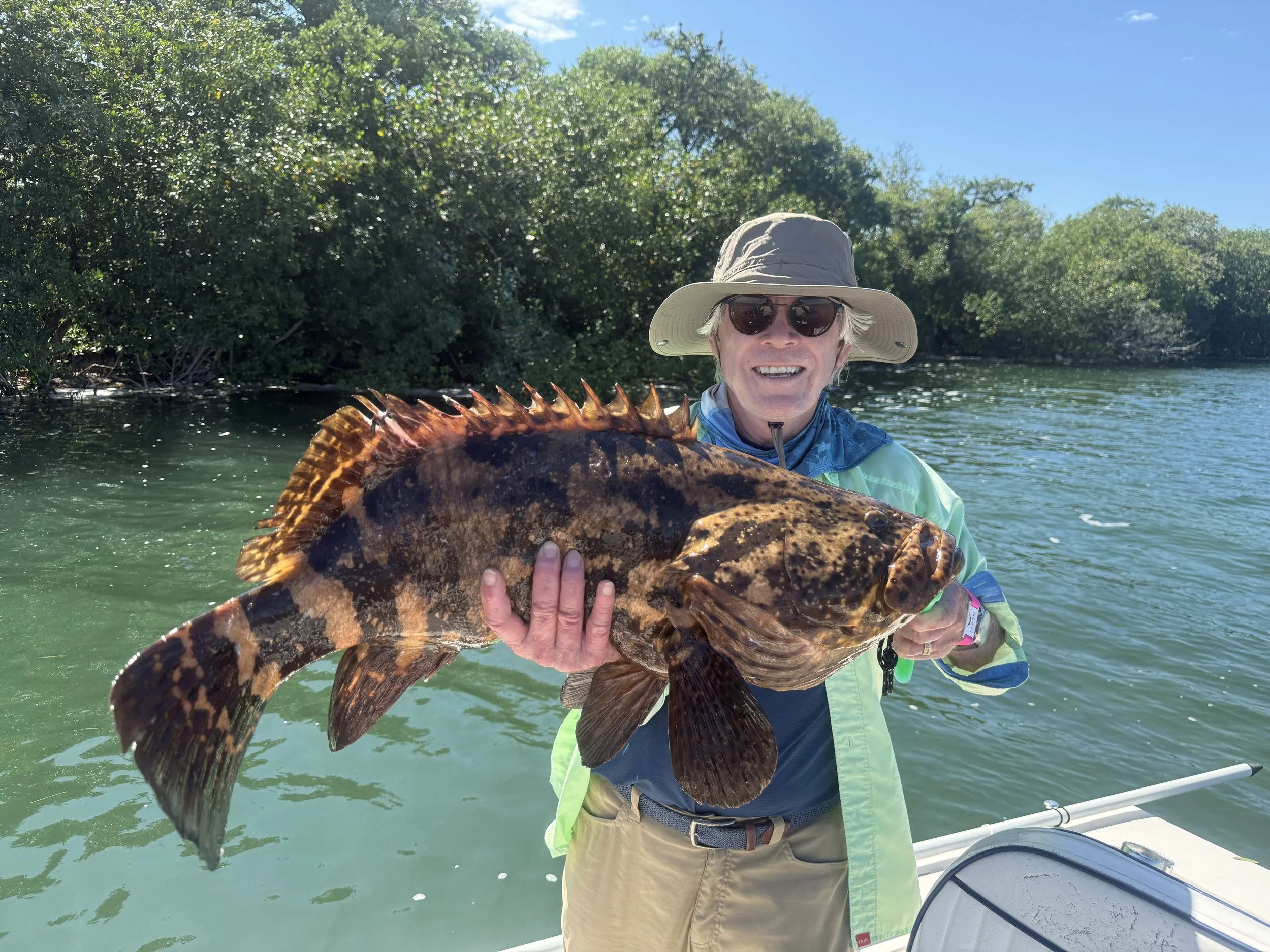An older man wearing a wide-brimmed hat, sunglasses, and a light jacket on a boat, holding a large fish with dark brown and orange coloring, spines along its back, and a wide mouth, with a backdrop of water and green trees.