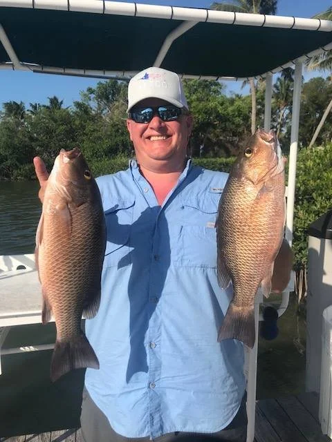 Person holding two large fish, wearing sunglasses, a white cap, and a blue shirt, standing on a boat with water and trees in the background.