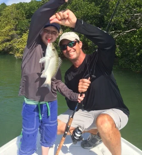 A young boy and Capt Tony Horsley on a boat, holding a fishing rod and showing off a fish they caught together, with green water and trees in the background.