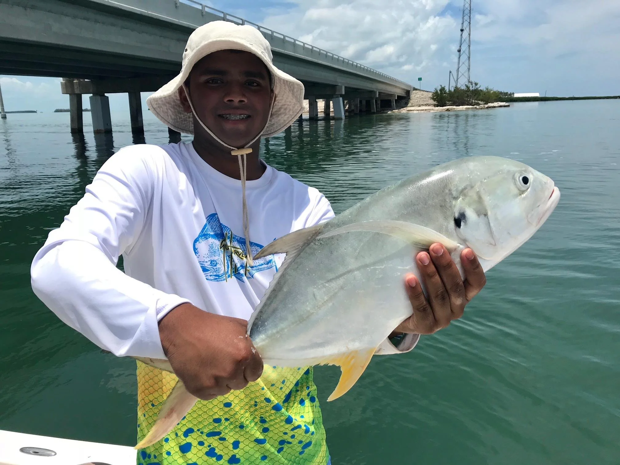 A young man wearing a wide-brimmed hat and a white shirt holding a large fish on a boat near a bridge over water.