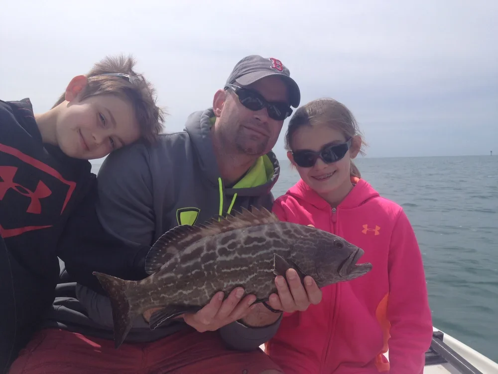 A man and two children on a boat holding a large fish they caught, with the ocean in the background.
