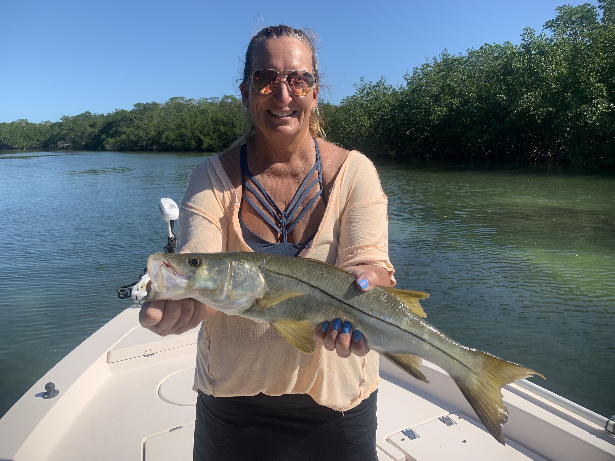 A woman wearing sunglasses and a beige top holding a large fish on a boat with water and green trees in the background.