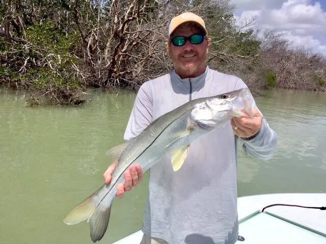 Man in sunglasses and cap holding a large fish on a boat in a river with trees in the background.