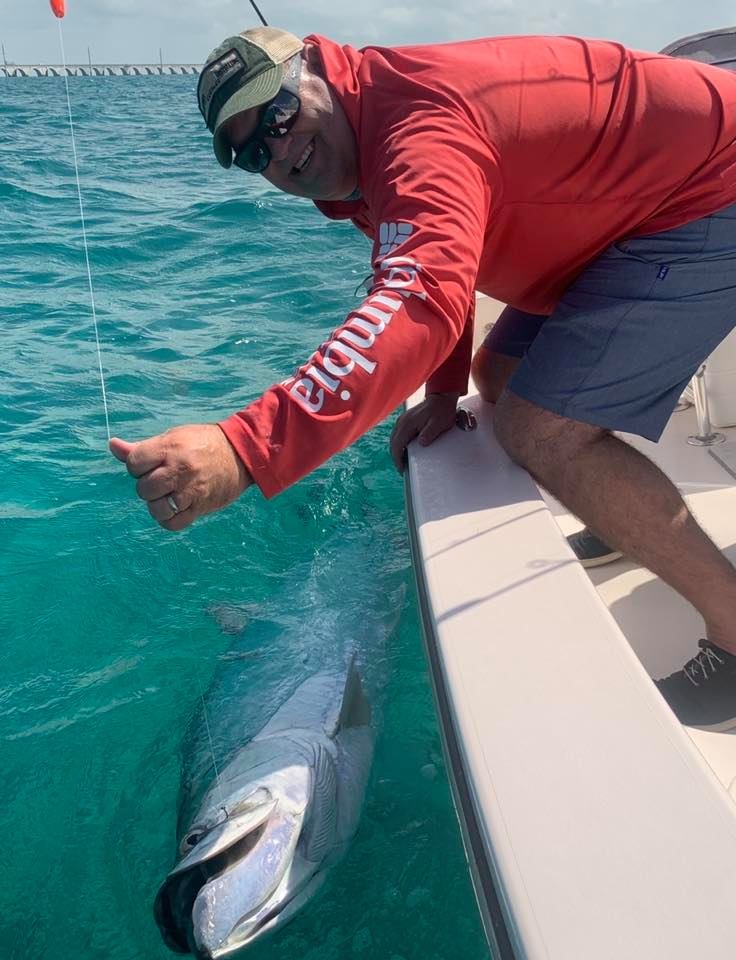 A man in a red jacket, gray shorts, and a cap is on a boat, giving a thumbs-up while fishing near a large shark in clear blue water. The shark is close to the boat's edge.
