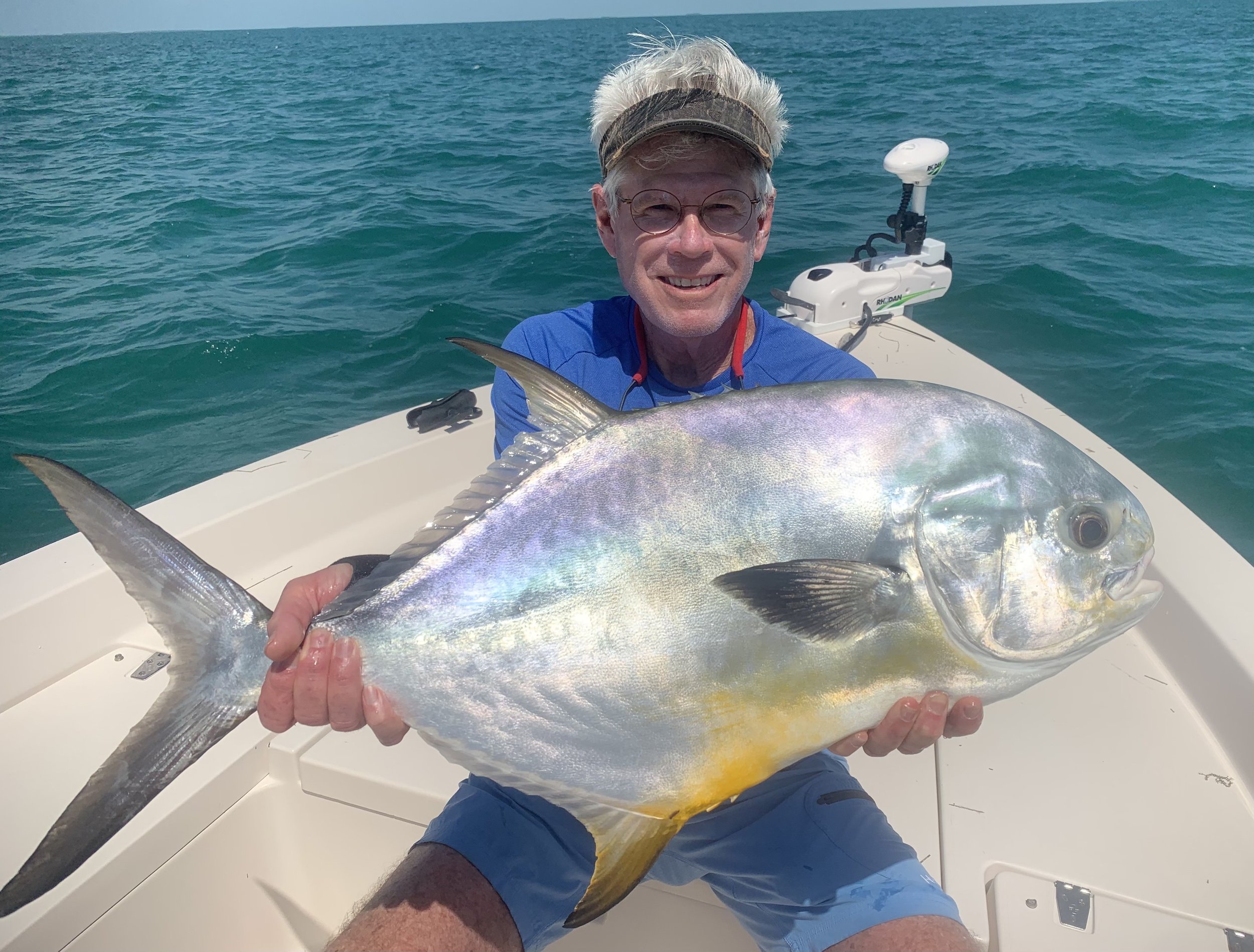 A man on a boat holding a large fish with shiny, iridescent scales and a yellow patch near its tail, in a blue ocean.
