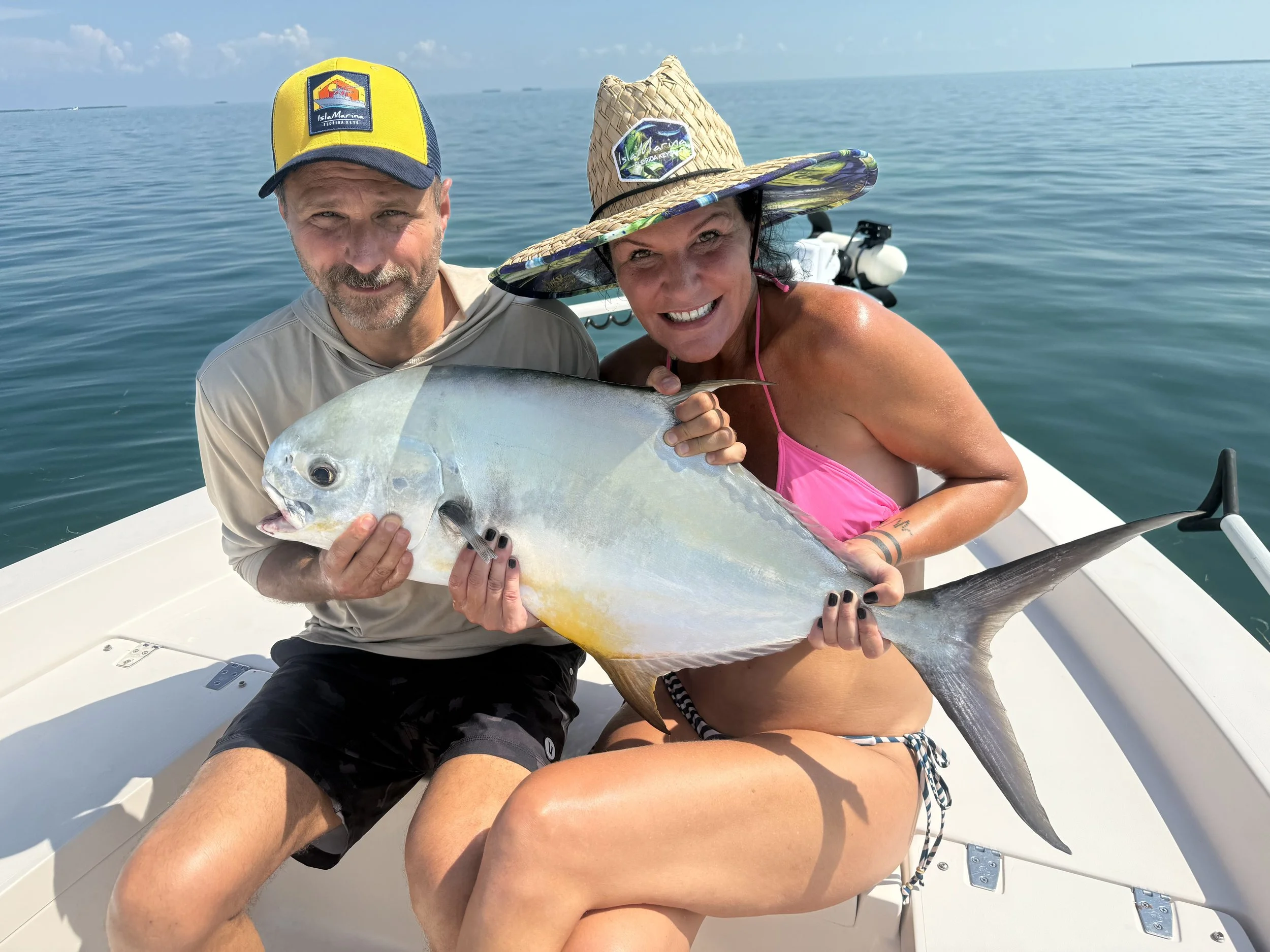 A man and woman on a boat, holding a large fish with an open mouth, a silvery body, and a long tail, smiling at the camera in an ocean setting.