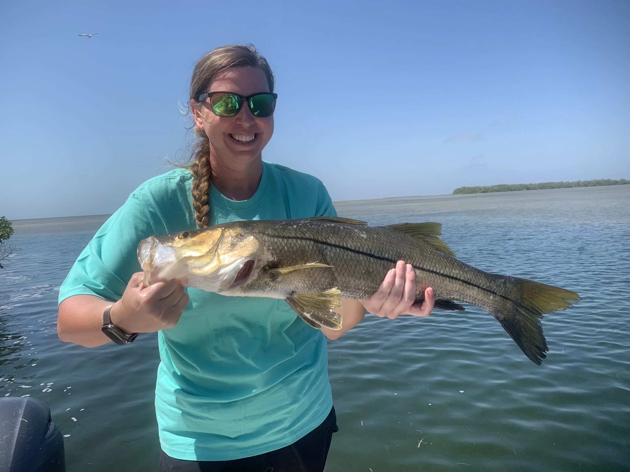 A smiling woman wearing sunglasses holding a large fish she caught, outdoors on a body of water with clear skies.