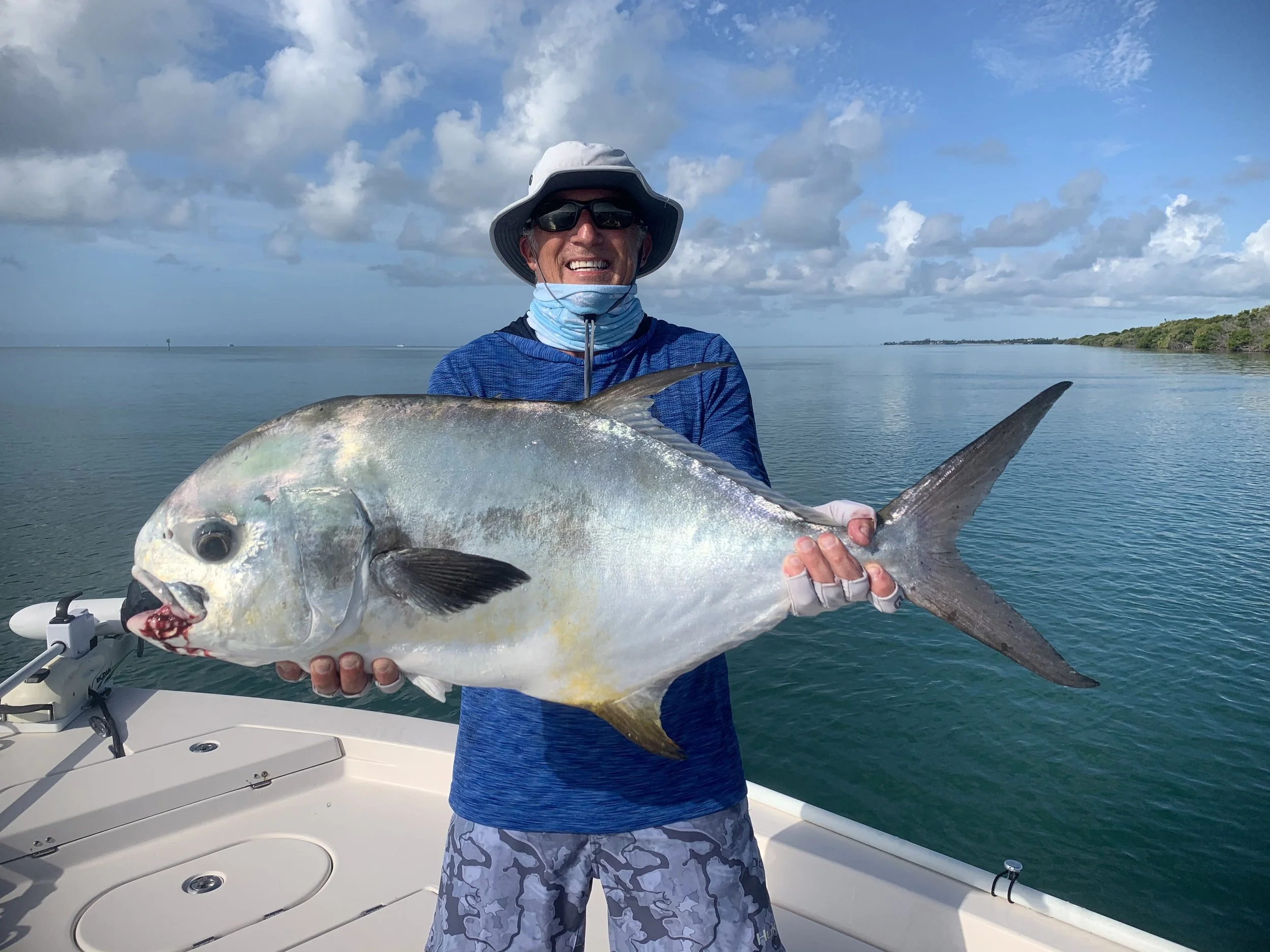 A man on a boat holding a large fish, smiling. The background shows calm water, a cloudy sky, and a distant shoreline.