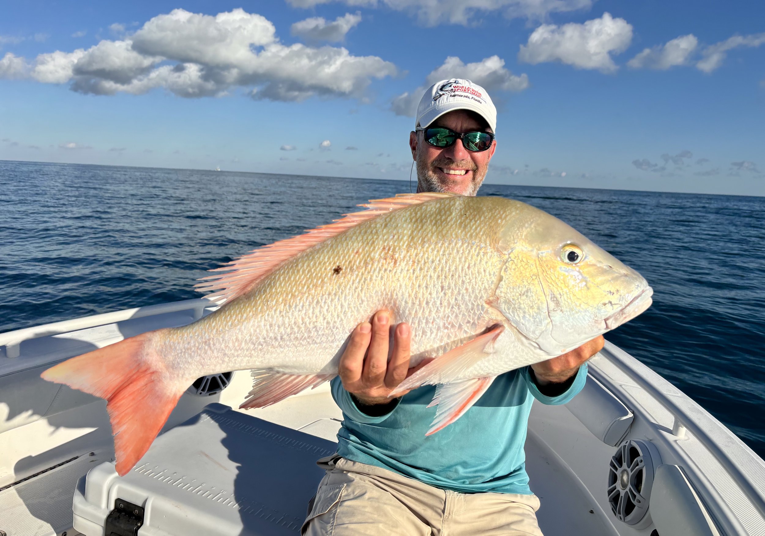 A man wearing sunglasses and a white cap holding a large fish on a boat in the ocean under a partly cloudy sky.