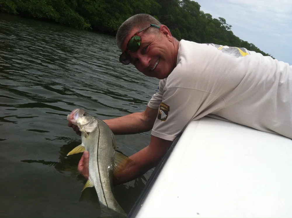 Man lying on a boat holding a fishing catch, smiling, with water and trees in the background.