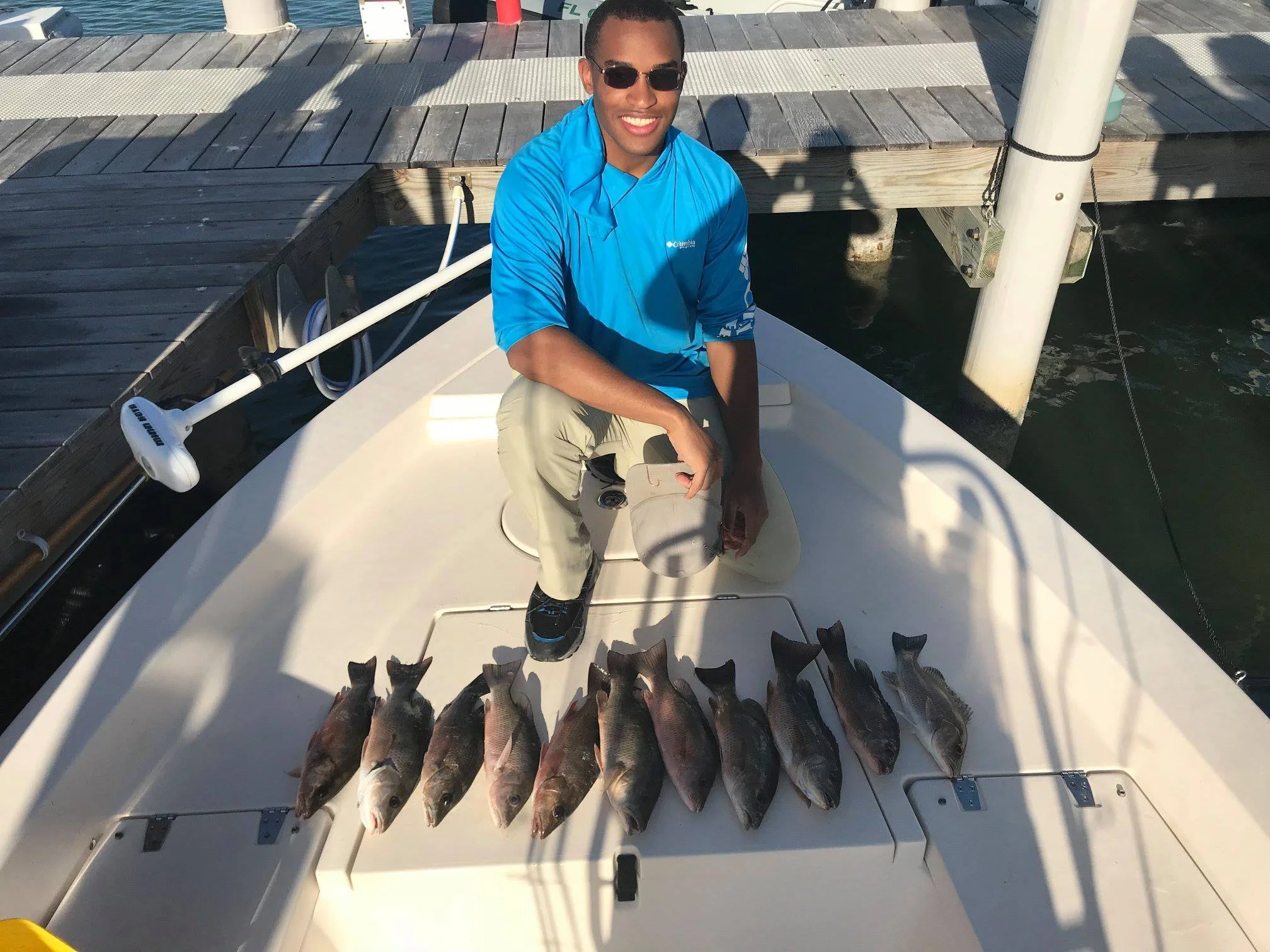 A man in a bright blue shirt and beige pants is sitting on a boat, smiling at the camera, with fishing rods nearby. In front of him on the boat deck are eight freshly caught fish lined up in a row.