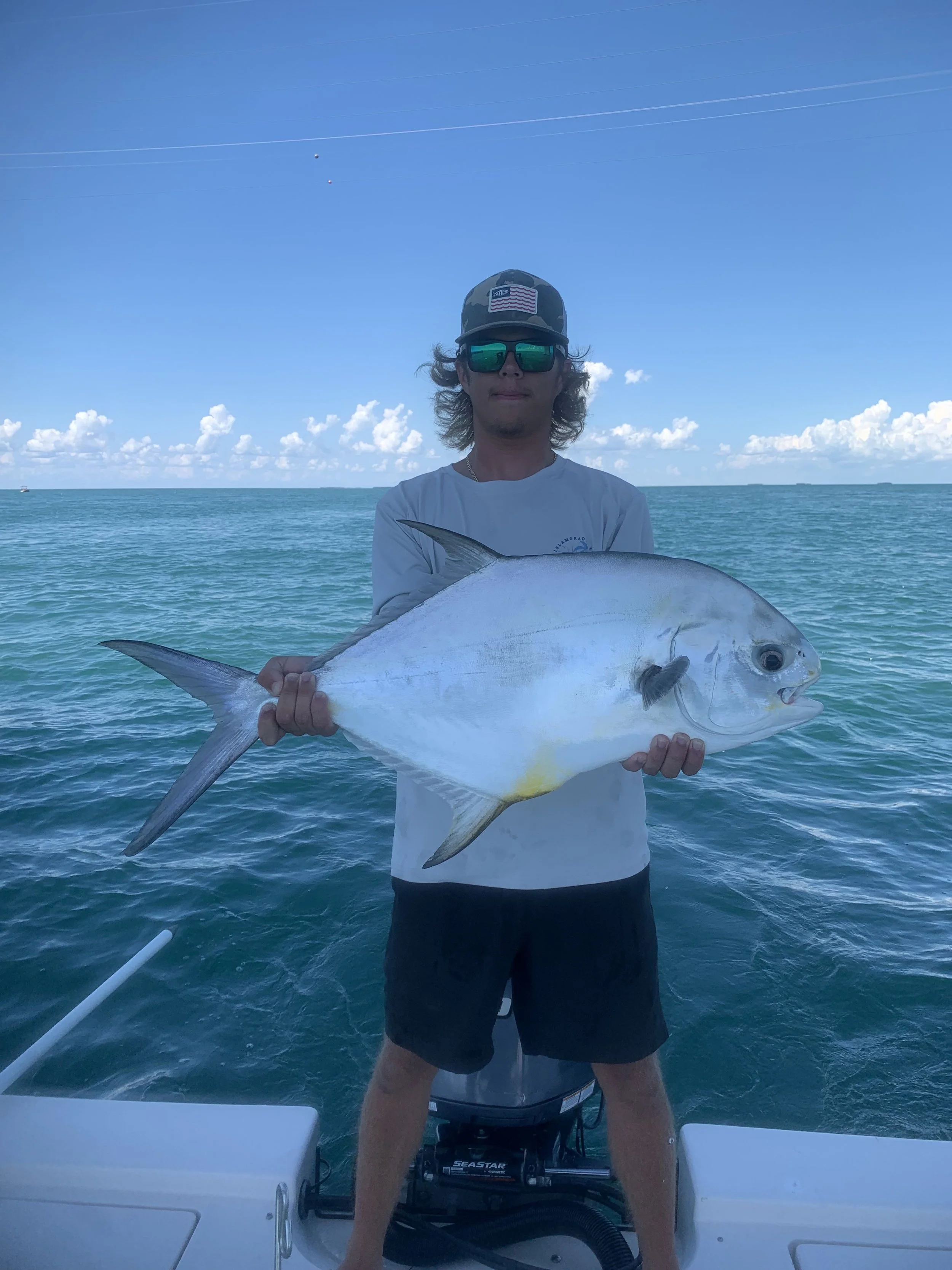 Man standing on a boat holding a large fish, with the ocean and blue sky in the background.
