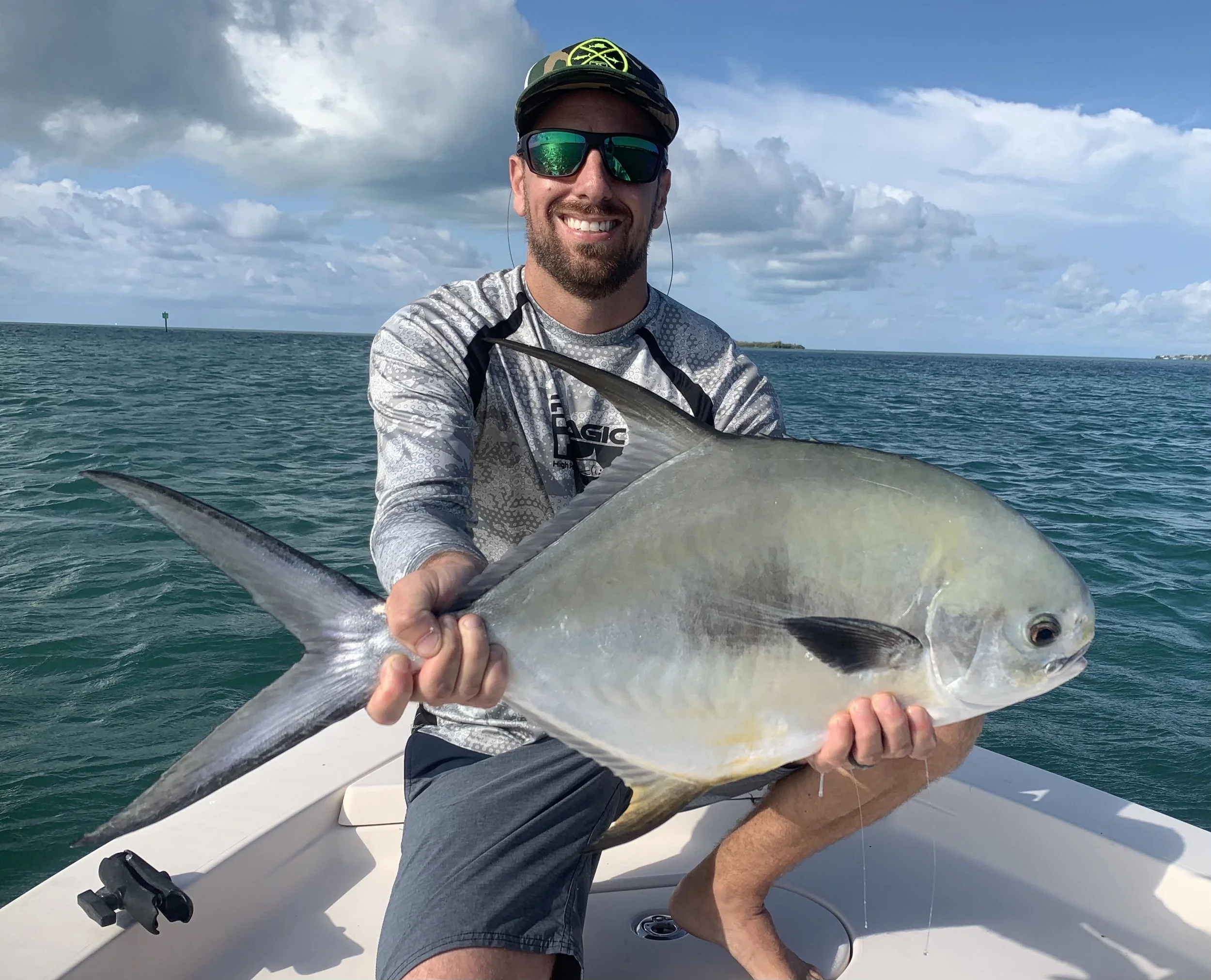 A man in sunglasses and a long sleeve shirt smiling while holding a large fish on a boat in the ocean.