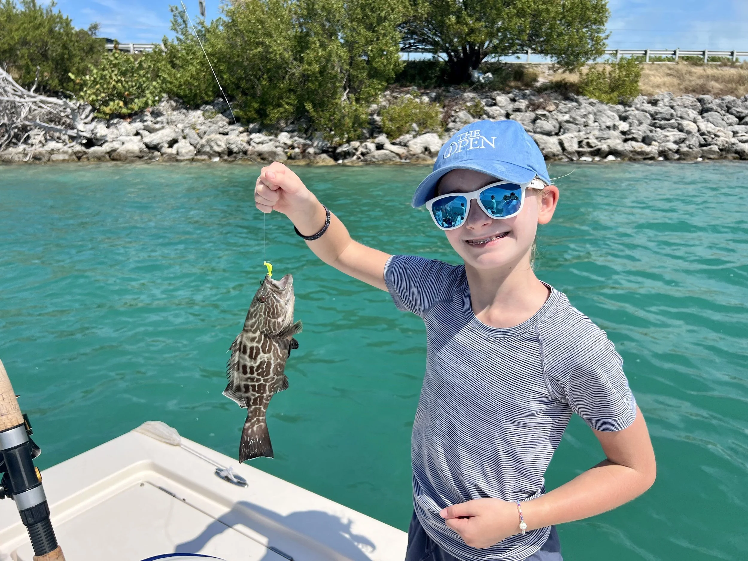 A young girl on a boat holding a fish she caught with a fishing hook, wearing sunglasses and a blue cap that says 'The Open', smiling, with water and rocky shoreline in the background.