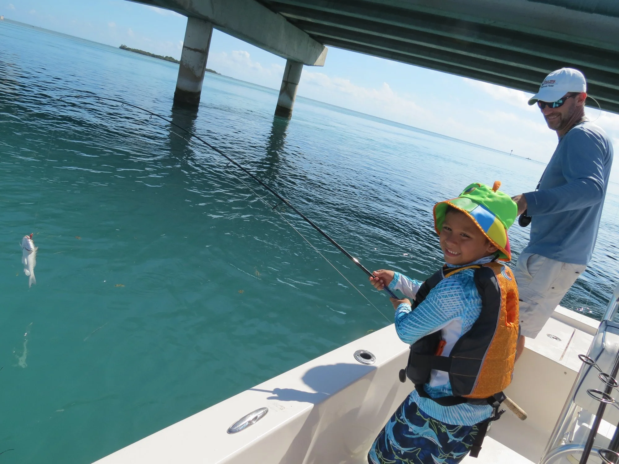 A man and a boy fishing from a boat under a bridge, with the boy holding a fishing rod and a fish.