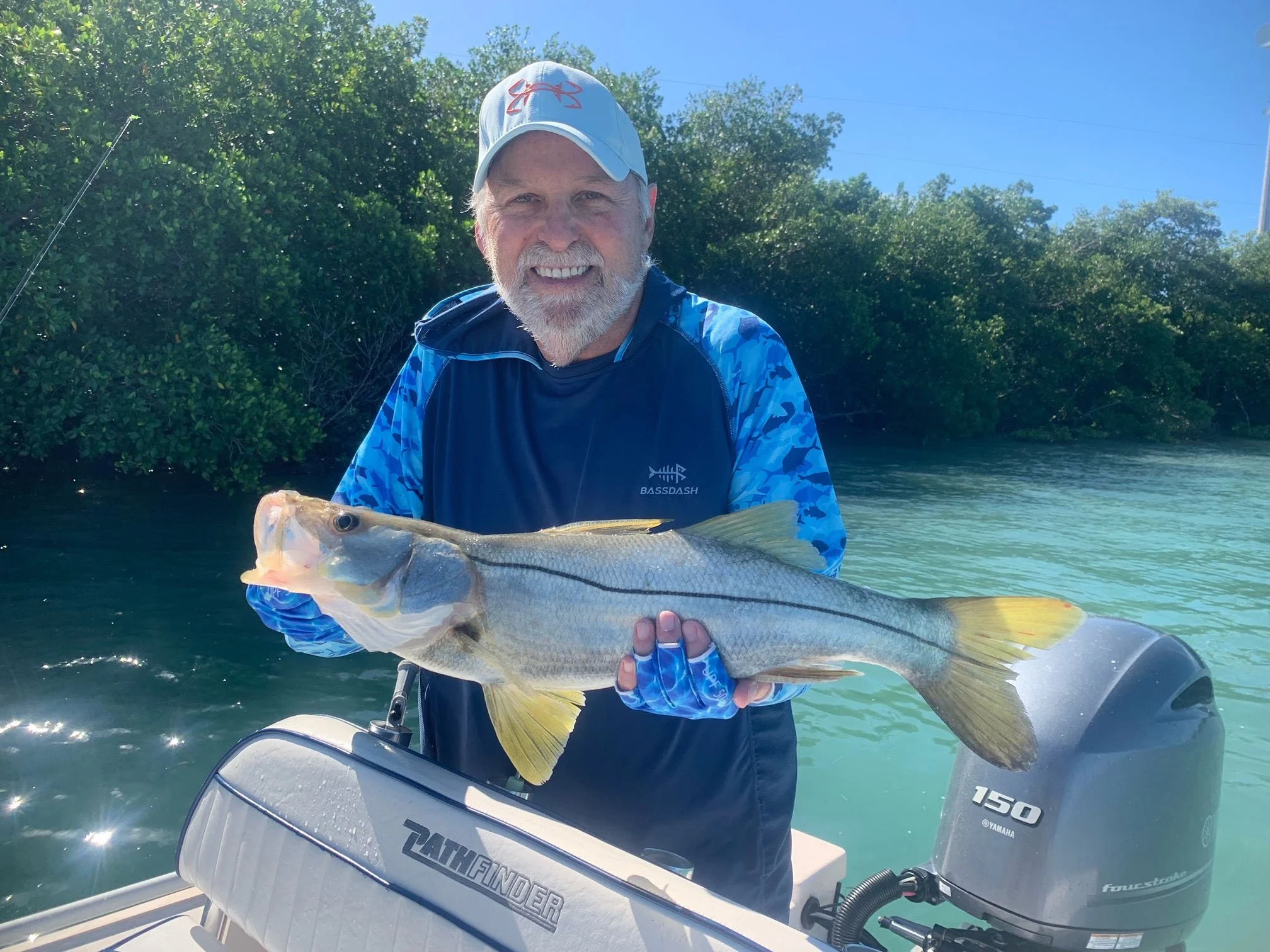 A man with a gray beard and wearing a cap and blue fishing shirt, smiling, holding a large fish on a boat with a Yamaha 150 outboard motor, with water and greenery in the background.