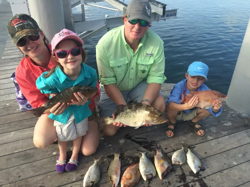 A family of four showing off their caught fish on a dock by the water. The mother, father, and two children are kneeling or squatting with the fish in their hands, with additional fish laid out on the dock.