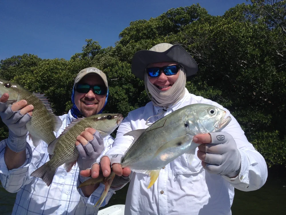 Two men in outdoor fishing gear holding caught fish on a sunny day with green trees in the background.