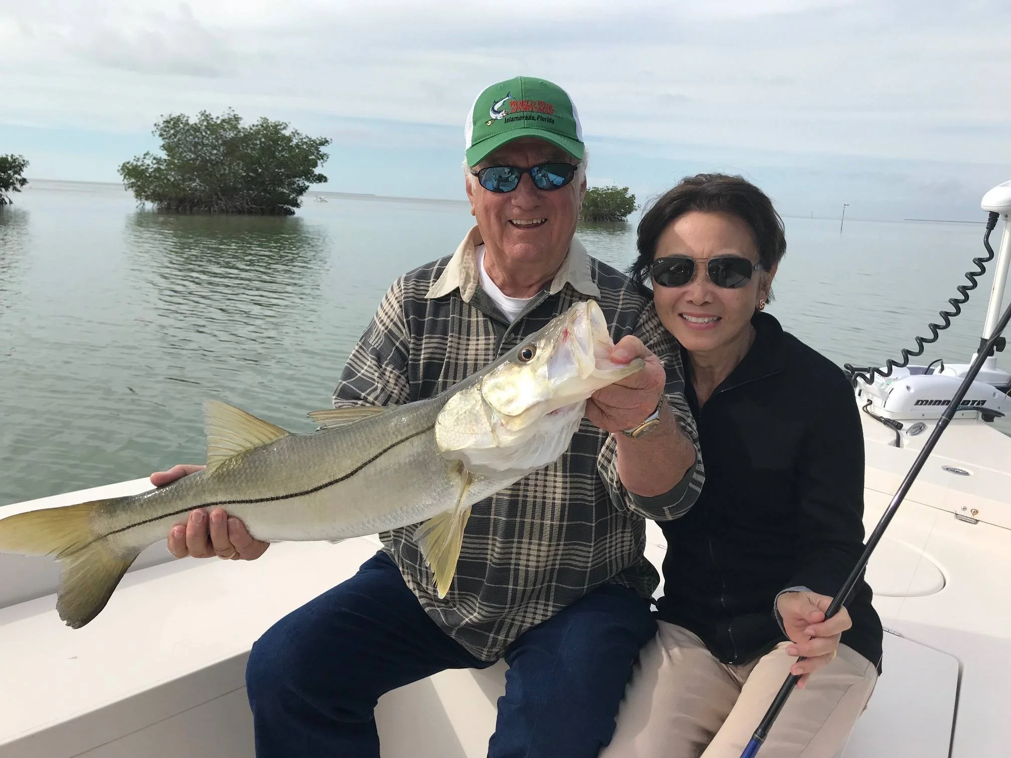 An older man and woman sitting on a boat, holding a large fish they caught together, with water and trees in the background.