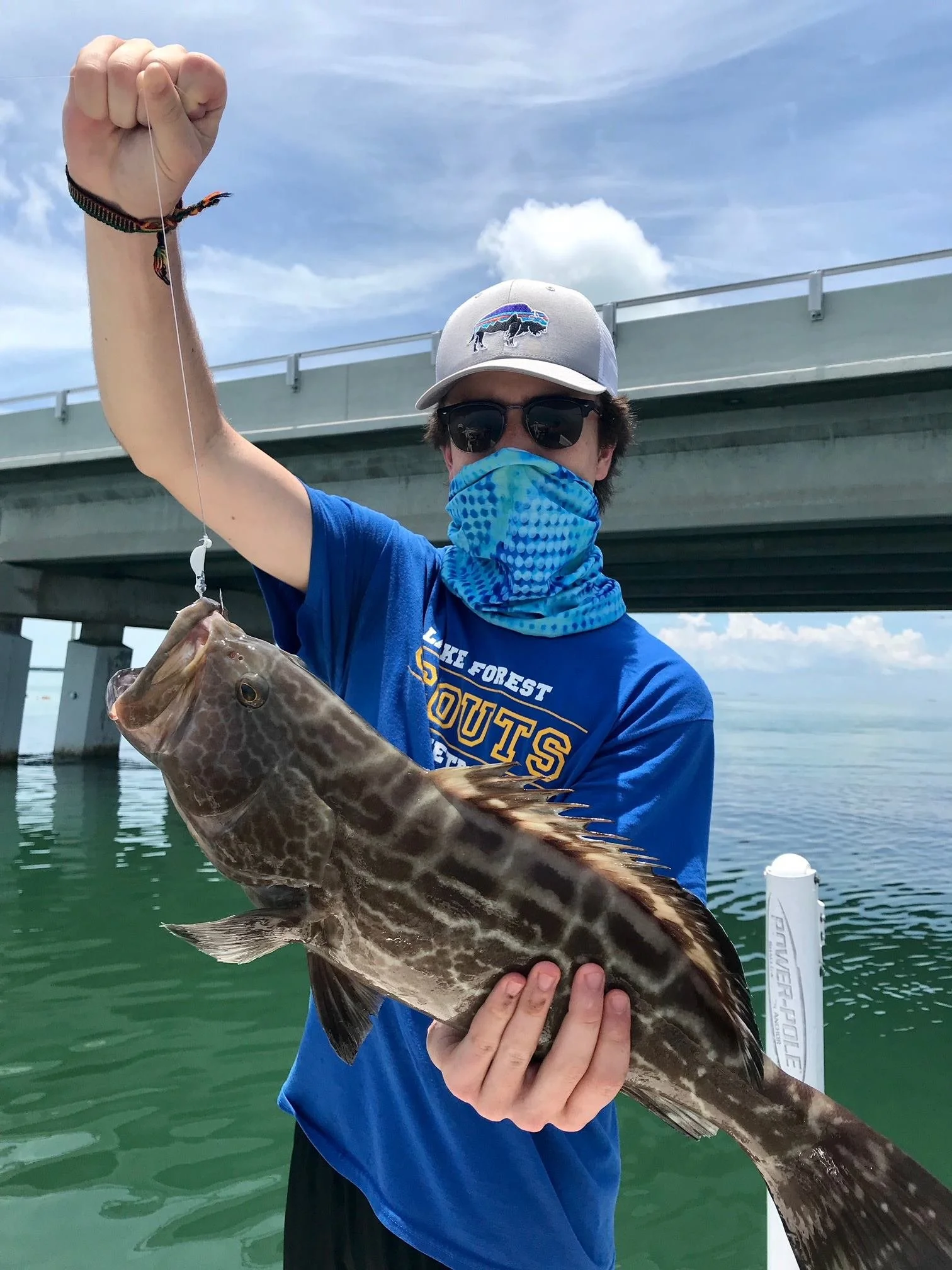 Person wearing a blue shirt, hat, sunglasses, and face mask holding a large fish with a bridge and cloudy sky in the background.