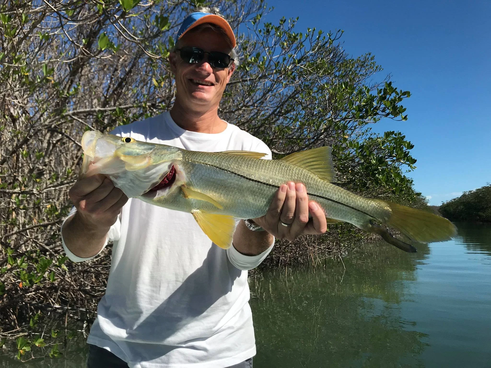 A man wearing sunglasses, a white long-sleeve shirt, and an orange cap holds a large fish in front of a water body with trees and bushes in the background.