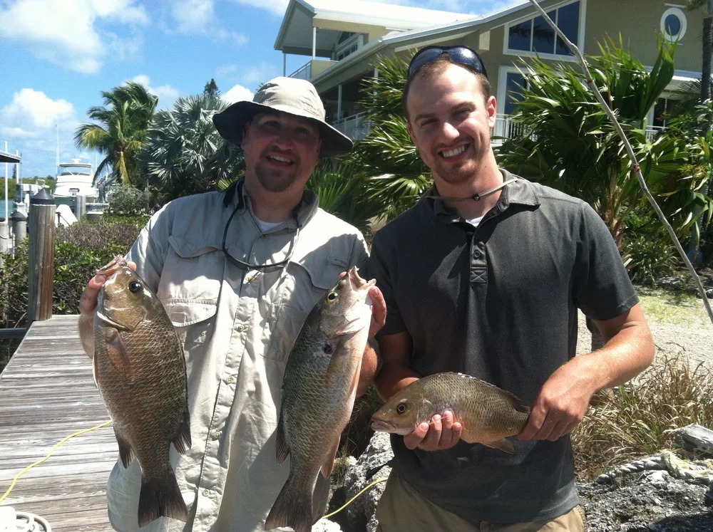 Two smiling men holding freshly caught fish outdoors near a dock with boat slips and lush green plants in the background.