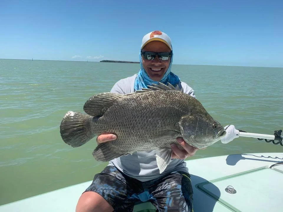 A smiling man in sunglasses, a white cap, and a blue neck gaiter holding a large fish on a boat in the water under a clear blue sky.